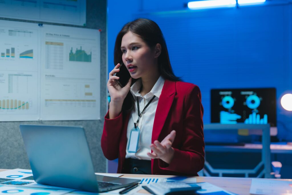 Businesswoman in a red jacket on the phone while working on a laptop with charts in the background | CMIT Solutions Cyber Security Business guide - Businesswoman-in-a-red-jacket-on-the-phone-while-working-on-a-laptop-with-charts-in-the-background